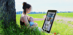 mamma med bäbis i knä framför ett träd på ett fält. Håller enorm iPad i handen.